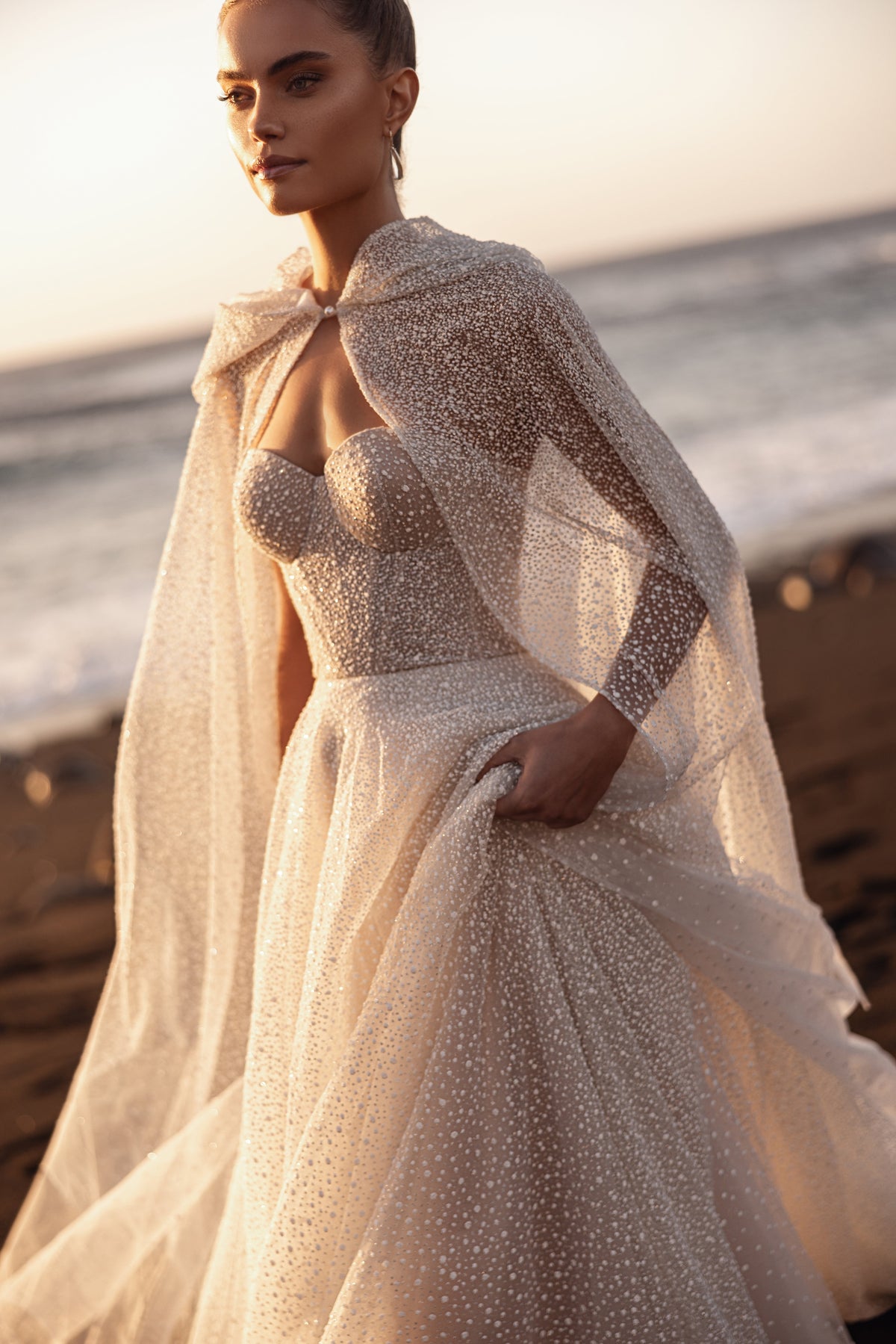 Amira on a beach, an open hooded cape over an A-line glitter tulle gown