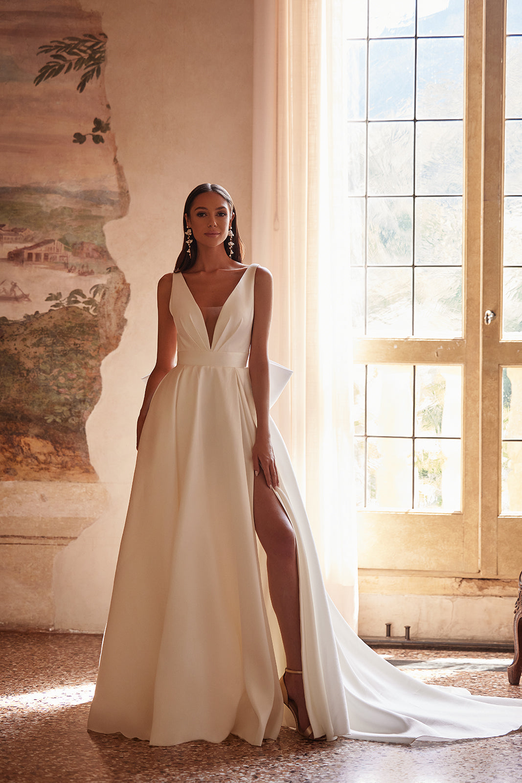 Full-length view of Margo wedding dress beside a window indoors, with a slit and train