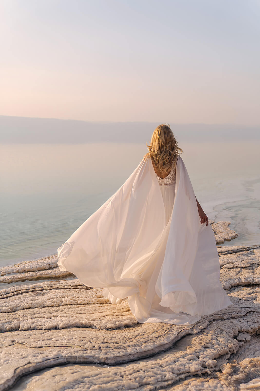 Full-length back view of Atena wedding dress, chiffon panels billowing above a salt shore