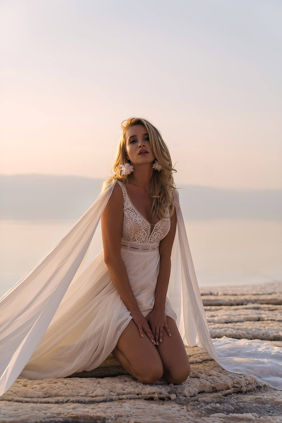 Bride kneeling on a salt shore in Atena wedding dress, lace bodice above spread chiffon panels