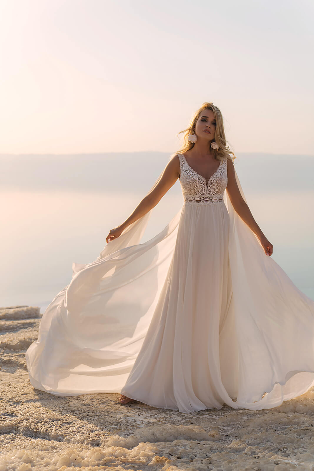 Front view of Atena wedding dress by the water, the bride holding its chiffon panels out