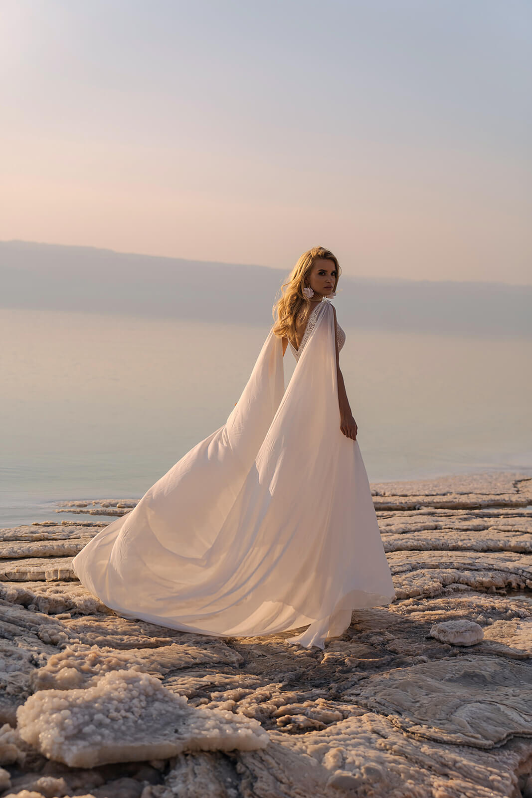Atena wedding dress from the side by the shore, with chiffon panels blowing from the shoulders
