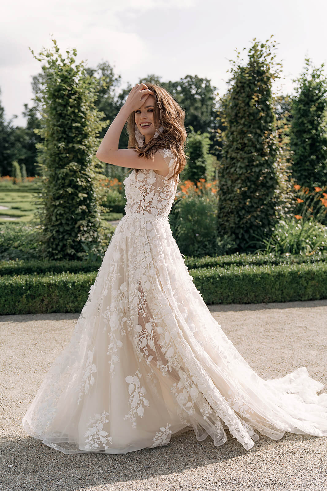 A bride walking along a garden path in Azalea wedding dress, floral lace running its full length