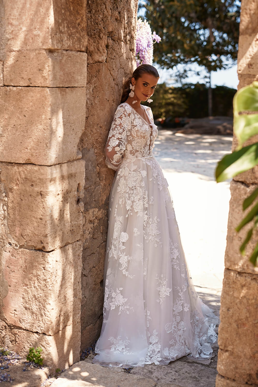 Full-length side view of Asami A-line wedding dress by a stone wall, with a lace train
