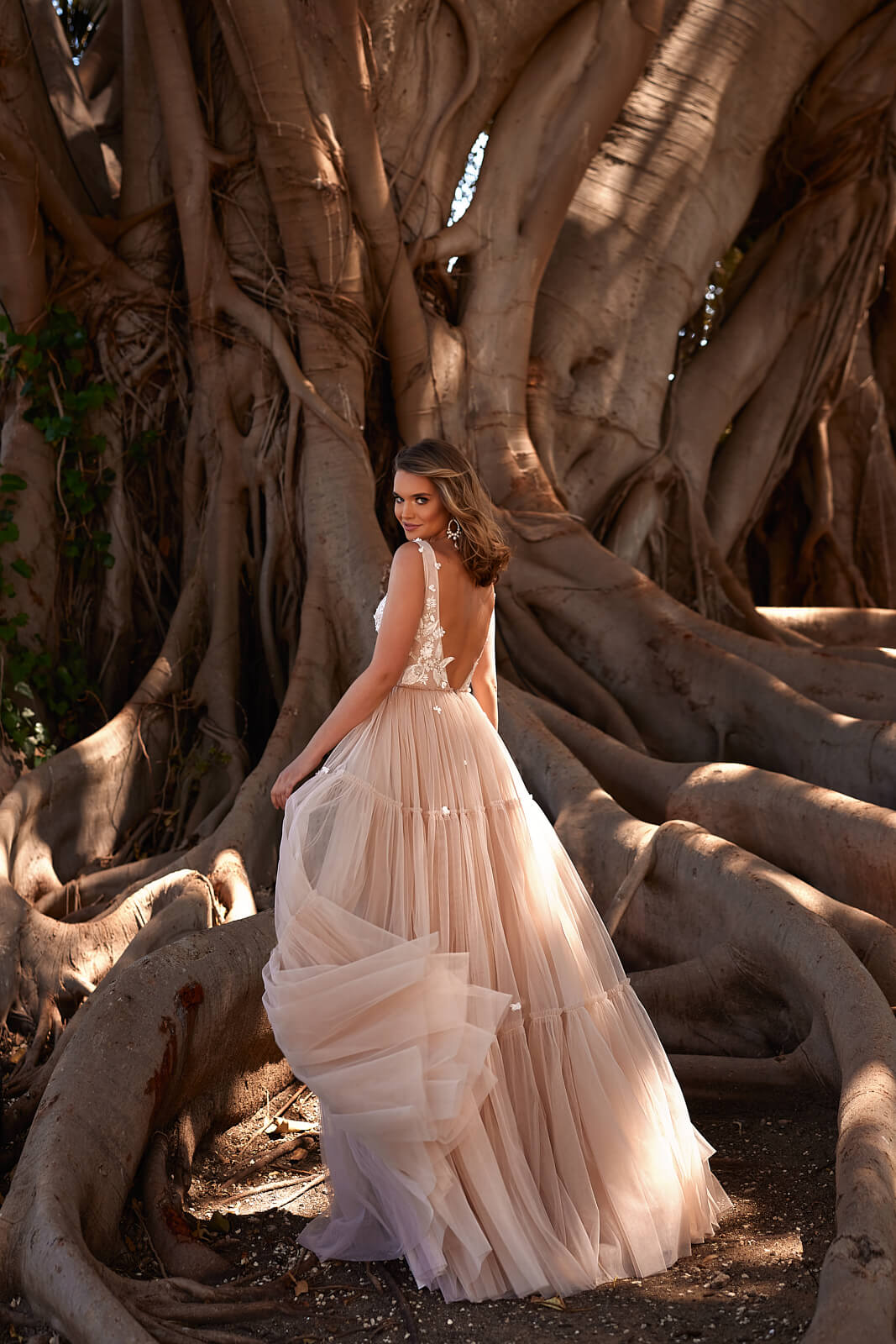 Full-length back view of Vanessa beside a tree, open back above the lifted tulle skirt