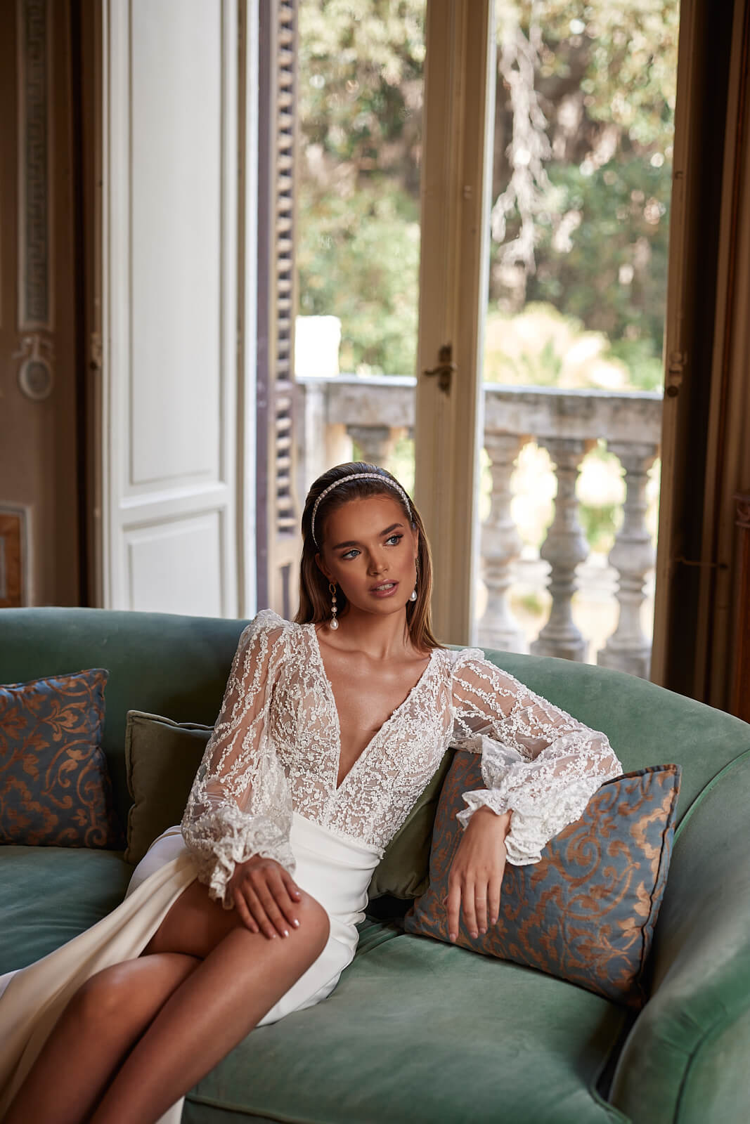 Bride seated on a sofa in Paula, showing lace sleeves and a high skirt slit