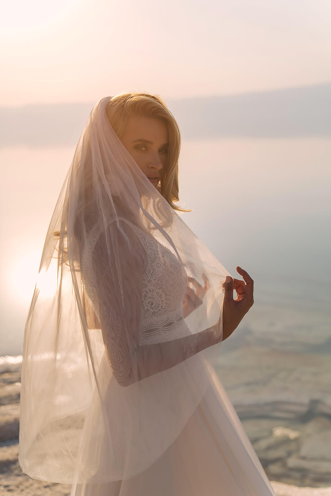 Regular veil against the light by the water, tulle drawn over the bride's face in profile