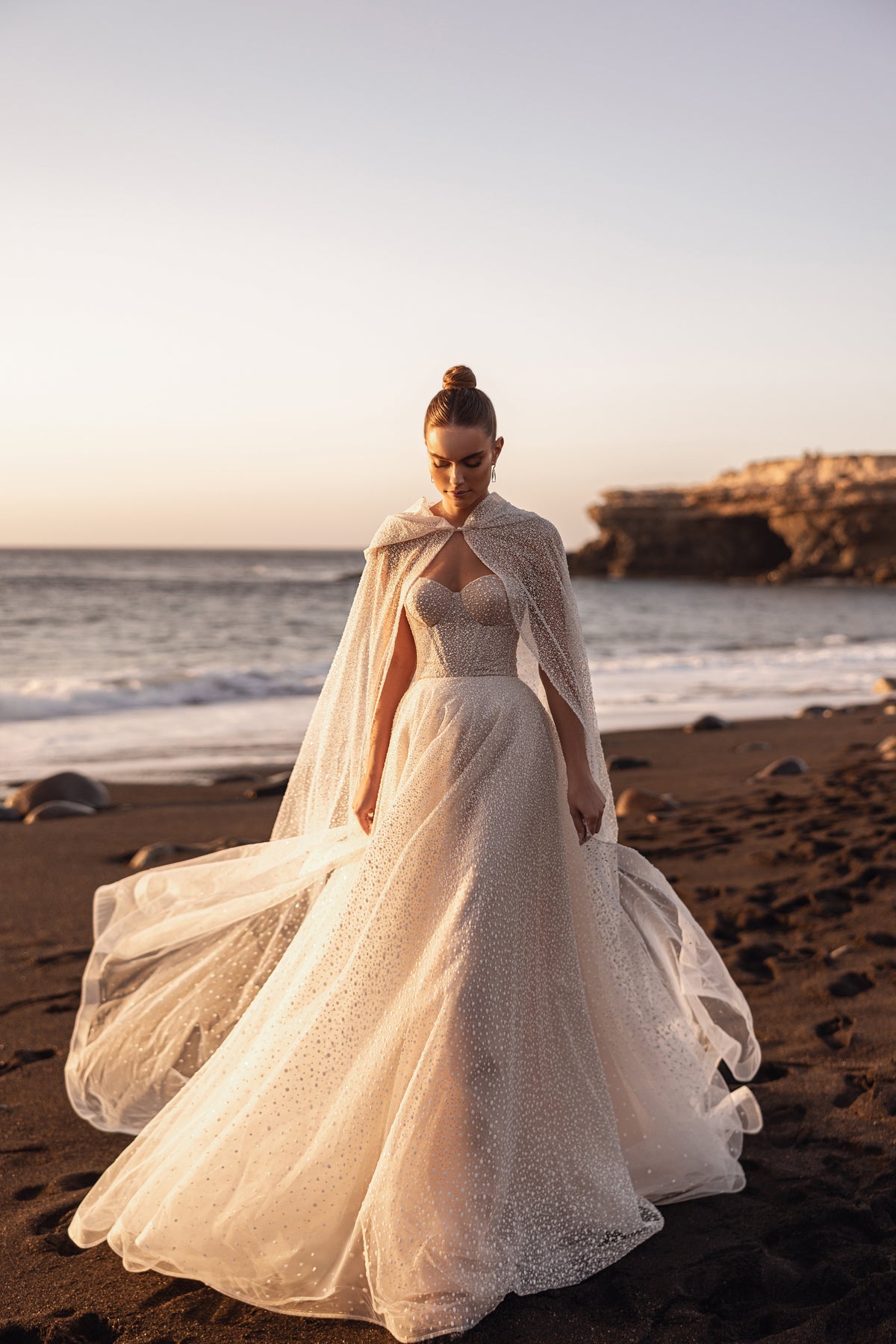 Full-length view of Amira cape spread wide over a corset wedding dress on the beach