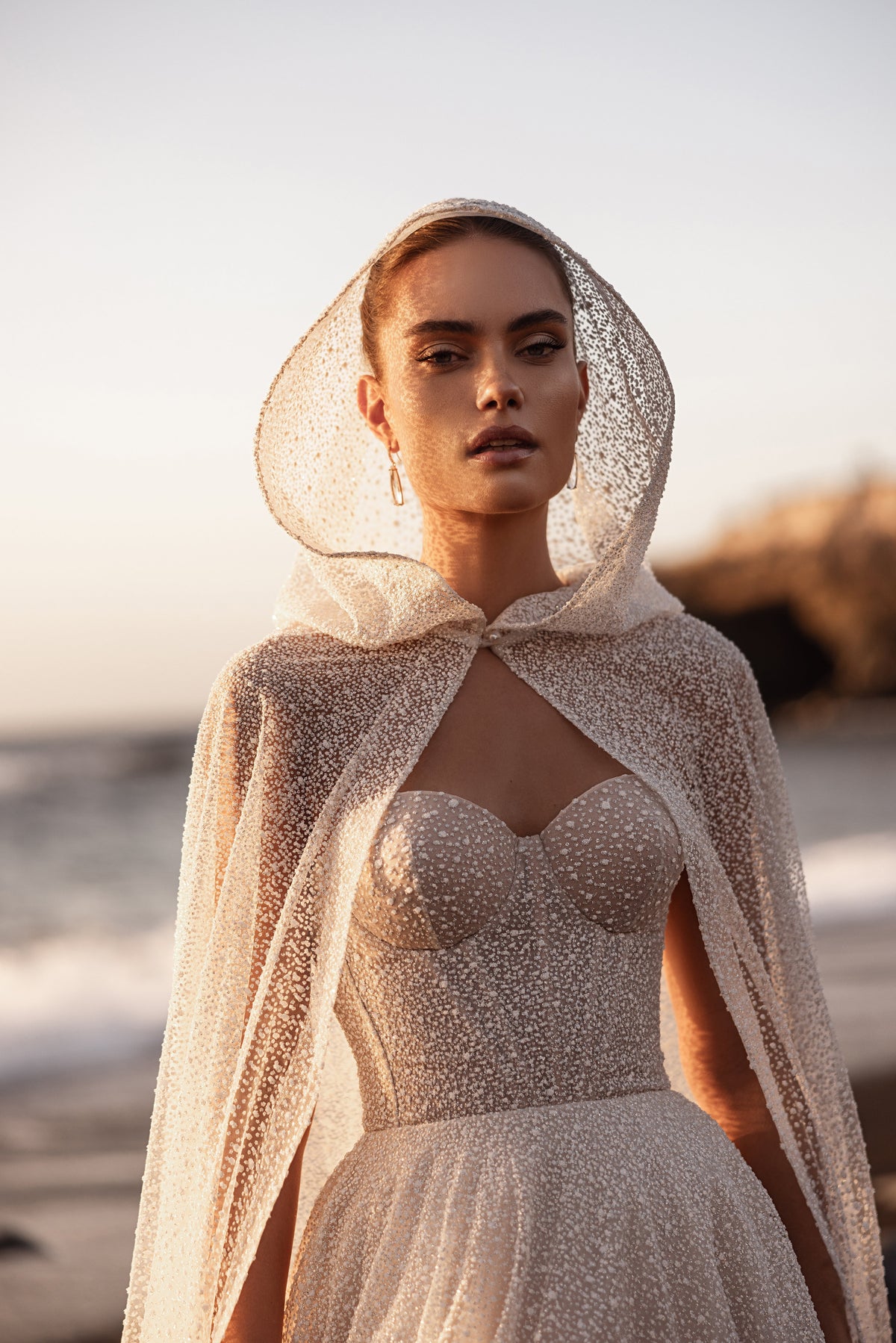 Waist-up view of Amira cape by the sea, its glittering tulle hood draped above the bodice