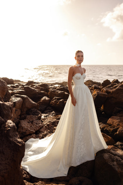 The Cholly bridal gown with a corset-style bodice and sweeping train, photographed at seaside sunset, evokes refined romance.