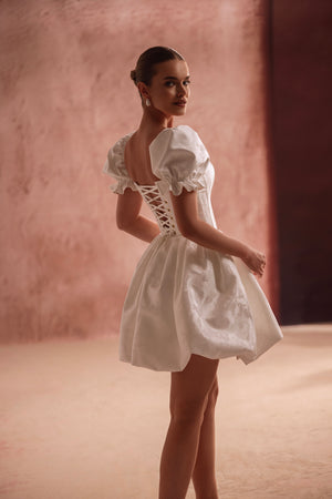 The Daphne bridal mini gown features a lace-up back and puffed sleeves, emphasizing a feminine and playful silhouette.