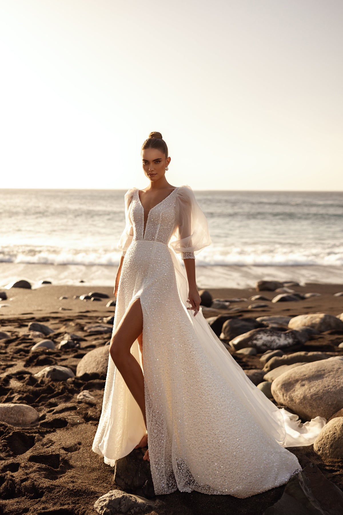 Full-length Davina wedding dress by the sea at sunset, with a high slit in the skirt