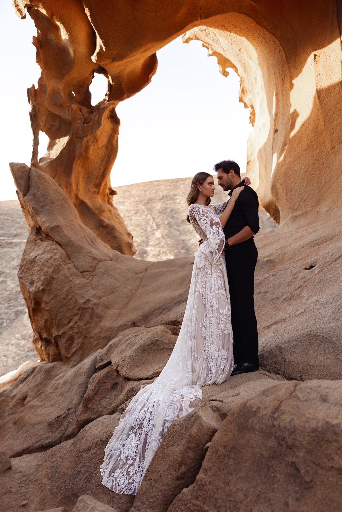 Full-length Dune lace wedding dress beneath a rock arch, the bride and groom in the desert