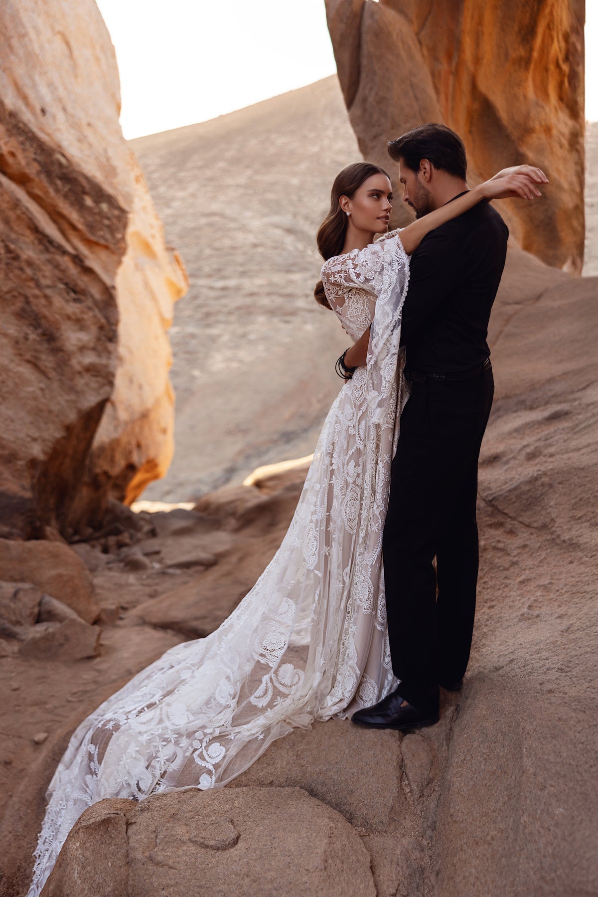 Dune wedding dress beside the groom on desert rocks, with a long lace train