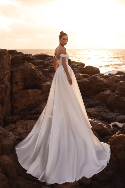 Mellisandre off-the-shoulder wedding dress with a dramatic train glowing in sunset light, perfect for beach or outdoor weddings.