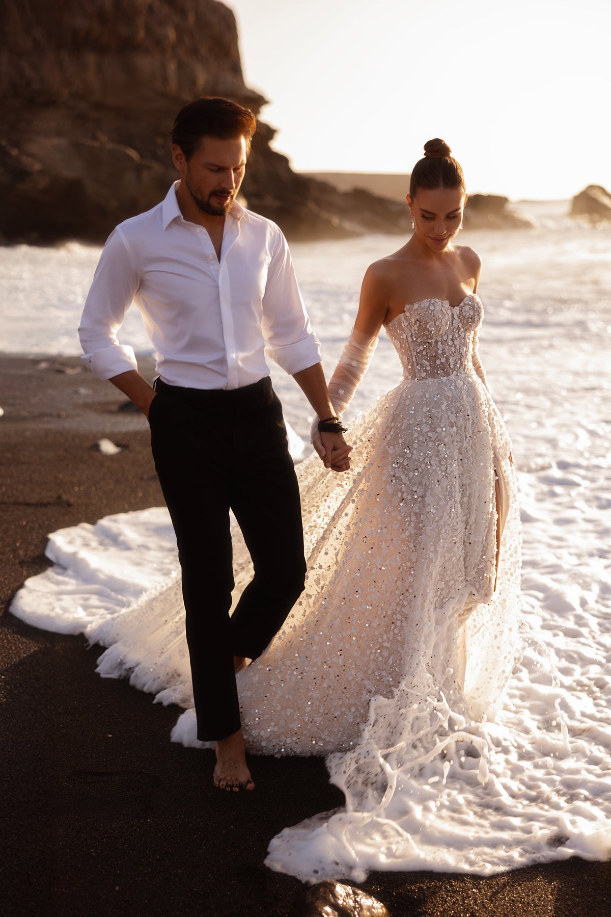 Bride and groom walking by the sea, the train of Reem wedding dress trailing in the water