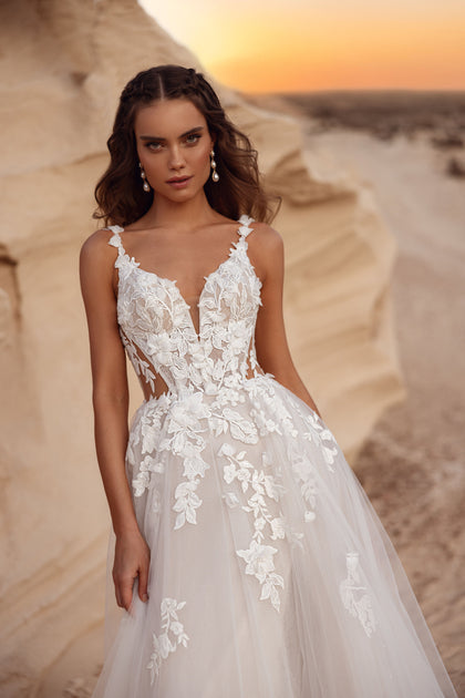 The Zafira bridal gown with a flowing tulle skirt and floral lace accents, styled with a groom in a beige suit, embodies timeless romance.