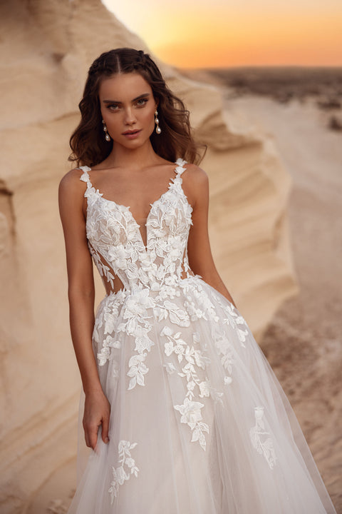 The Zafira bridal gown with a flowing tulle skirt and floral lace accents, styled with a groom in a beige suit, embodies timeless romance.